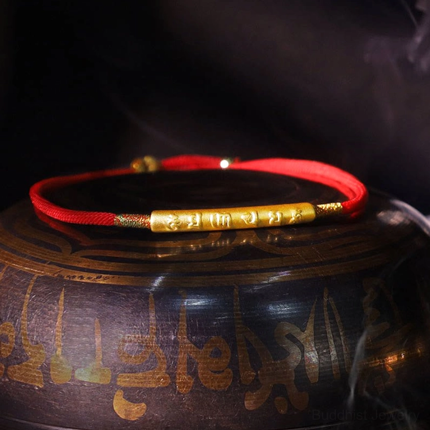 Om Mani Padme Hum red string bracelet with gold tube charm and metal bead details on Tibetan script vessel