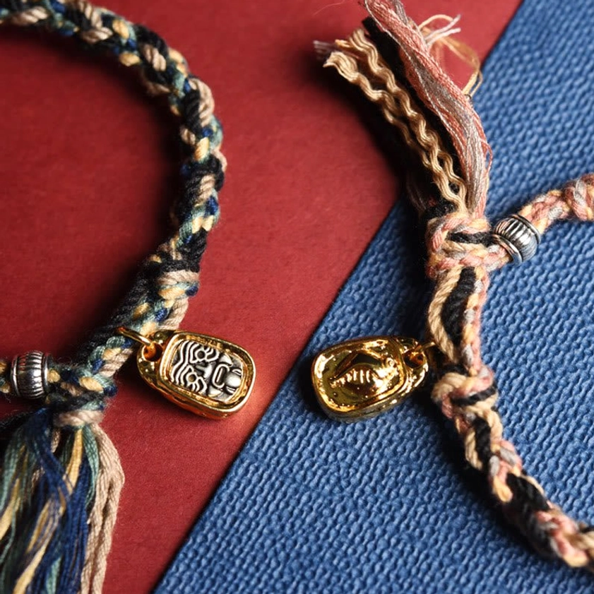 Two handmade Buddhist bracelets with mixed color woven ropes, metal face charms and tassel ends, flat lay display