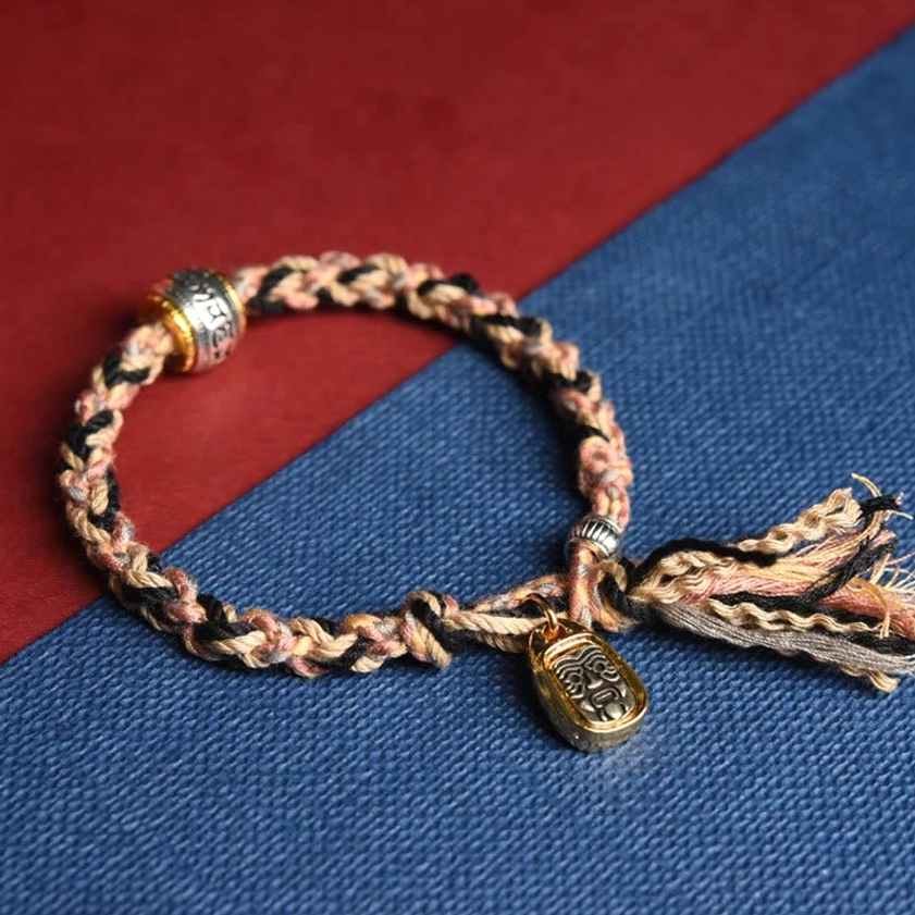 Pink beige black Buddhist bracelet with tan-brown woven cord, metal charm and tassel ends, close-up side view