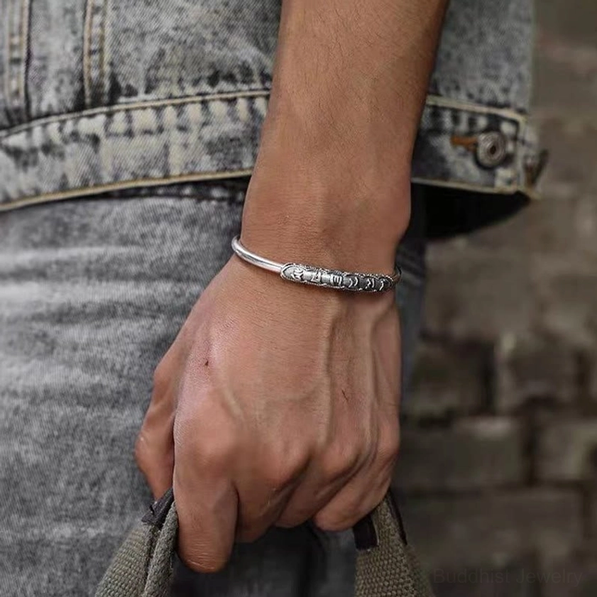 Om Mani Padme Hum engraved cuff bracelet worn on male wrist with casual denim outfit in outdoor brick wall setting
