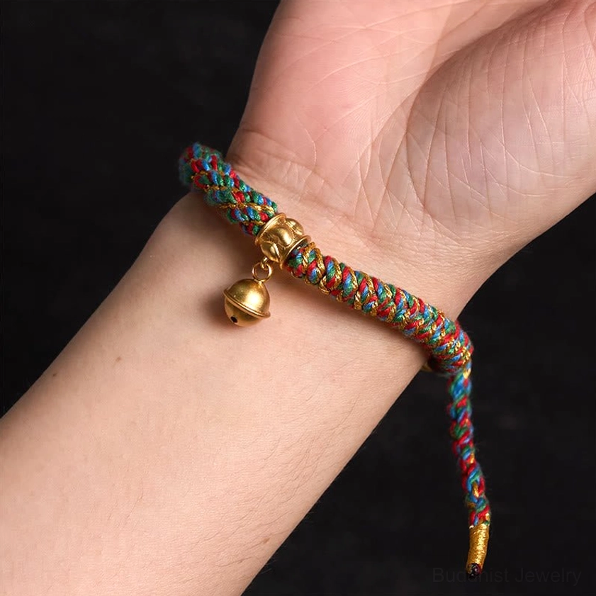 First-person wrist wear view of adjustable multicolor Buddhist string bracelet with gold bell pendant and tail cord