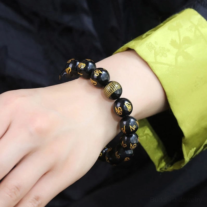 Wrist wearing mantra obsidian bracelet with golden Sanskrit characters on black beads, styled with traditional green fabric sleeve showing natural daily wear fit