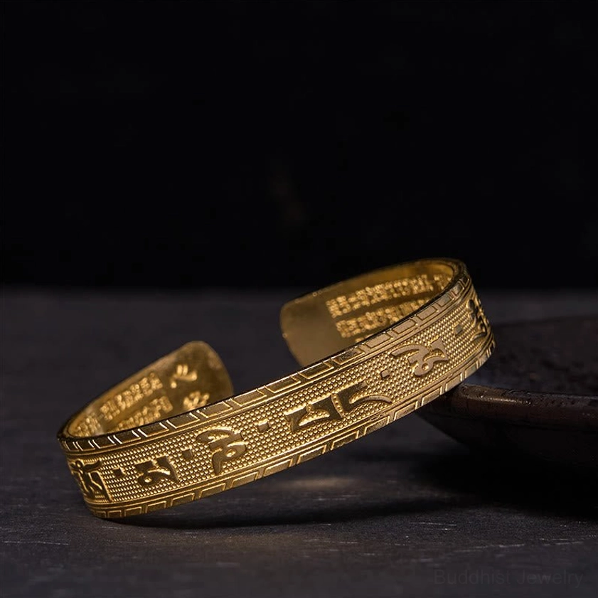 Polished brass Om Mani Padme Hum bangle with bright finish showing embossed six-syllable mantra, mirrored oblique overhead shot on dark wooden surface