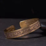 Om Mani Padme Hum brass bangle bracelet with antique bronze finish and embossed six-syllable mantra, viewed from oblique side angle on dark wooden surface