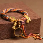 Om Mani Padme Hum Buddhist lucky string bracelet with multicolor braided cord and gold metal charms displayed on wooden box