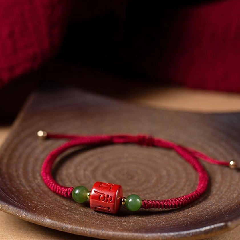 Om Mani Padme Hum cinnabar bracelet with braided rope and bead components on dark wooden tray with red fabric background