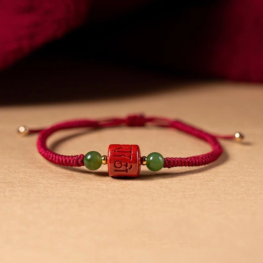Red braided rope Om Mani Padme Hum cinnabar blessing bracelet with engraved mantra bead and jade accents, top-down view on wooden table
