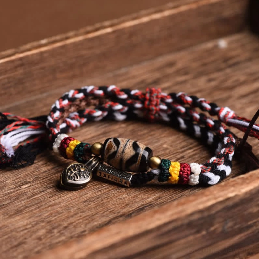 Double-wrap Tibetan Buddhist dzi bead bracelet with striped braided cord and metal charm tags on wooden surface