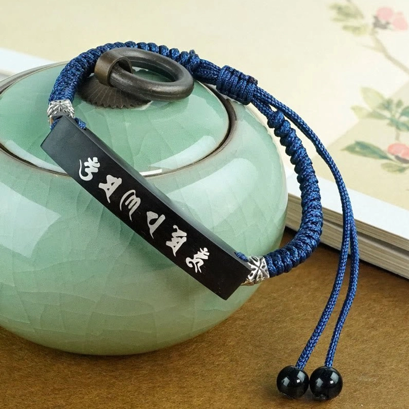 Silver-inlaid ebony mantra bracelet worn with green ceramic teapot and book, showcasing navy woven cord and adjustable design for Buddhist spiritual practice
