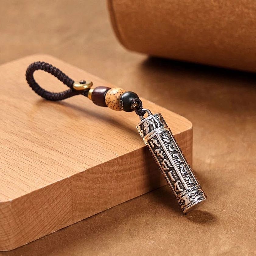 Buddhist mantra copper keychain side view displaying engraved cylindrical pendant with dark brown woven rope on wooden block background
