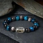 Silver Sheen Obsidian and Blue Tiger Eye Buddhist mantra bracelet with engraved Om Mani Padme Hum charm on slate stone backdrop
