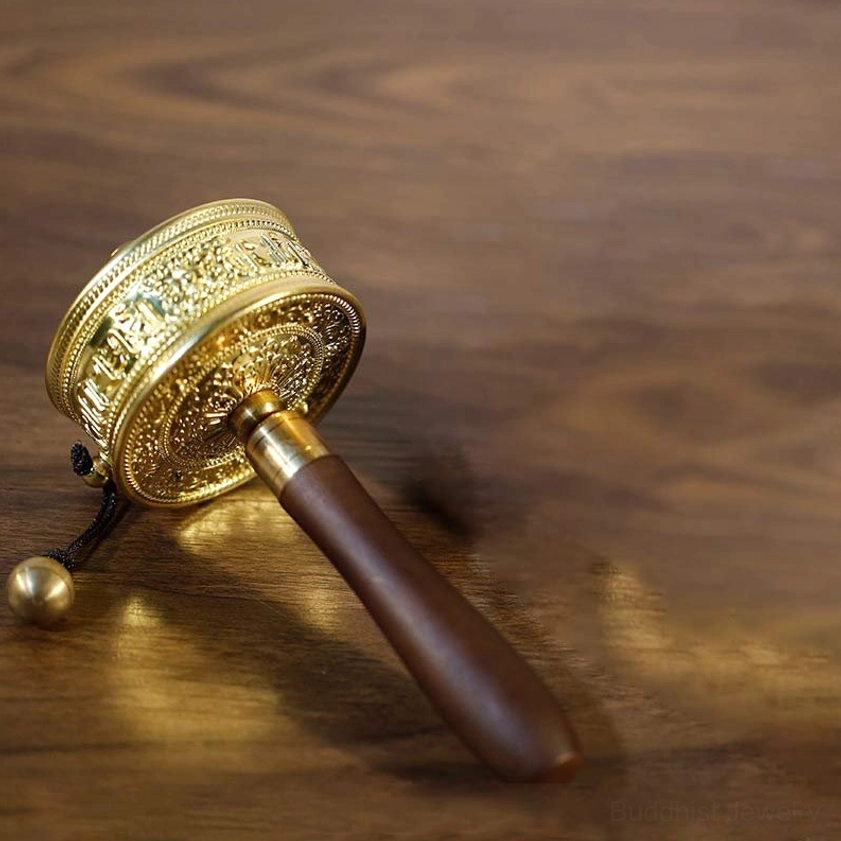 Buddhist prayer wheel spinning meditation tool, low angle view showing embossed gilt metal barrel, smooth sandalwood handle, and decorative hanging bell
