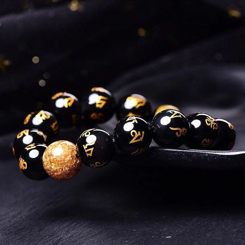 Sanskrit mantra obsidian bracelet three-quarter angle view displaying golden script on black beads and decorative gold foil accent bead on dark display tray