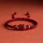 Red rope Om Mani Padme Hum cinnabar Buddhist bracelet with engraved mantra bead, gold spacers, and braided cord on terracotta background