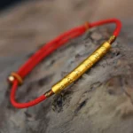 Om Mani Padme Hum red string bracelet with gold-plated sterling silver charm, overhead view on wooden texture background