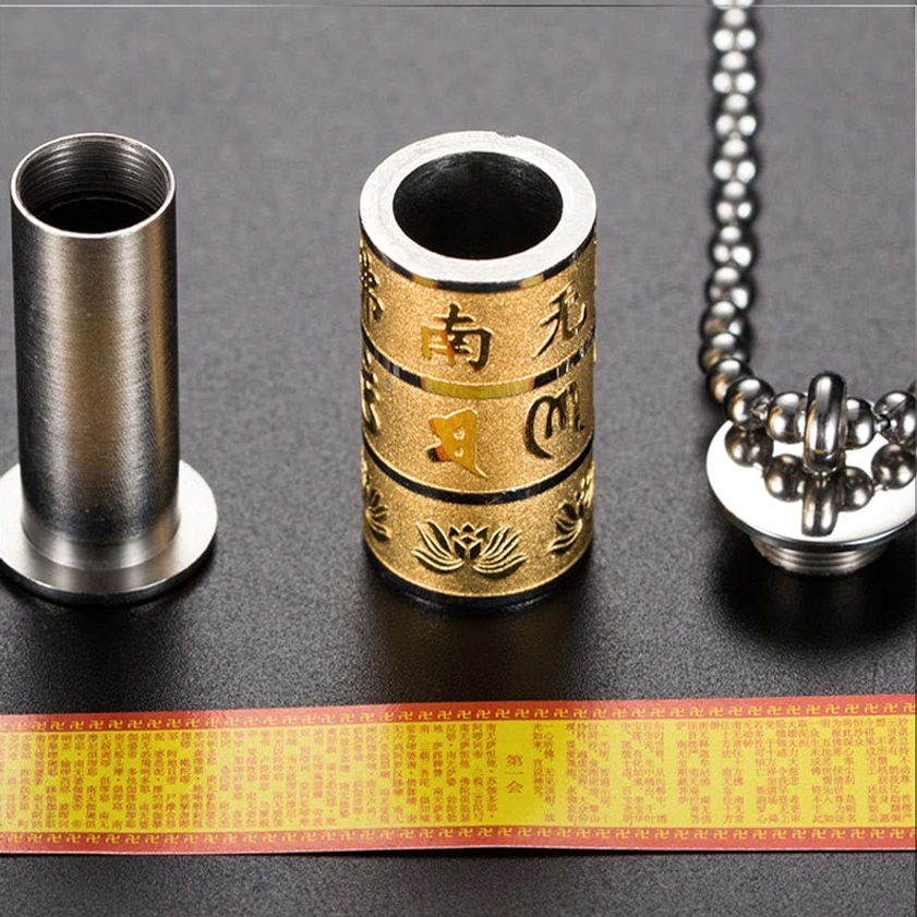 Removable cylindrical prayer wheel pendant with detachable chain showing hollow design, matte silver and gold finish with sacred Buddhist text engraving and lotus patterns, flat lay on black background