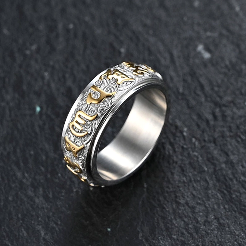 Six True Words Om Mani Padme Hum titanium steel spinner ring with silver-gold two-tone finish, embossed Sanskrit engravings, side view on dark stone background