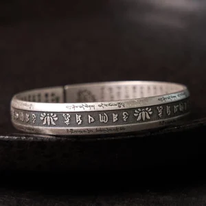 Om Mani Padme Hum silver cuff bracelet with engraved sacred text and lotus motif, top-down view on dark matte surface