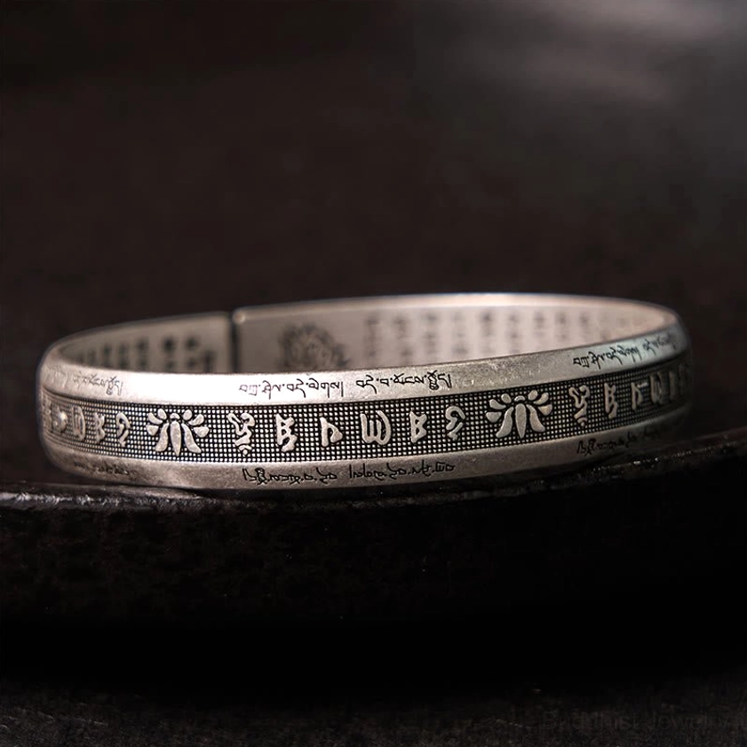 Om Mani Padme Hum silver cuff bracelet with engraved sacred text and lotus motif, top-down view on dark matte surface