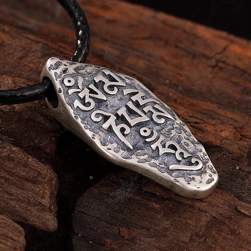 Buddhist mantra silver pendant showing right side angle view, carved Tibetan script on dark wooden board, matte silver tone finish