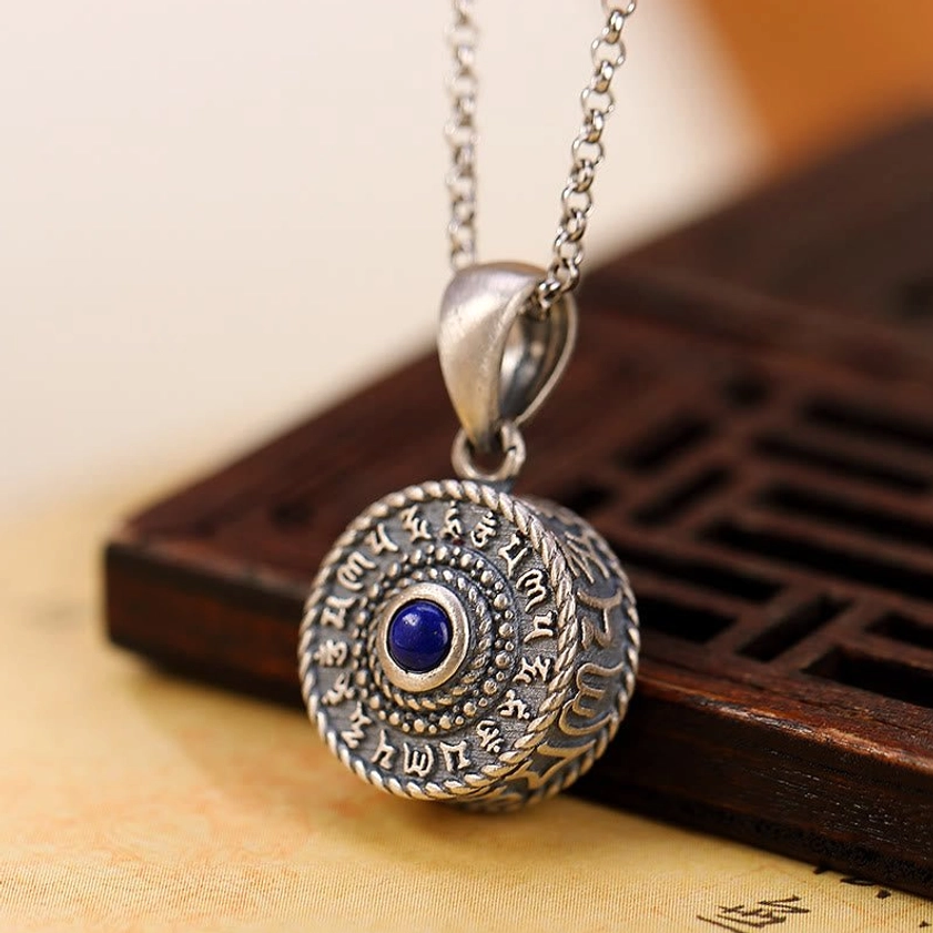 Three-quarter view of Om Mani Padme Hum silver pendant with lapis stone resting on wooden lattice and Buddhist scripture paper, showing sutra inscription