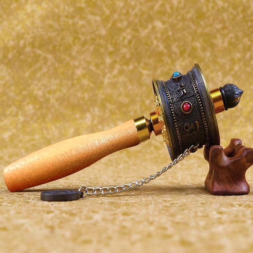 Buddhist prayer wheel with dark metal body featuring red and blue gemstone inlays, wood handle with hanging chain displayed on textile backdrop
