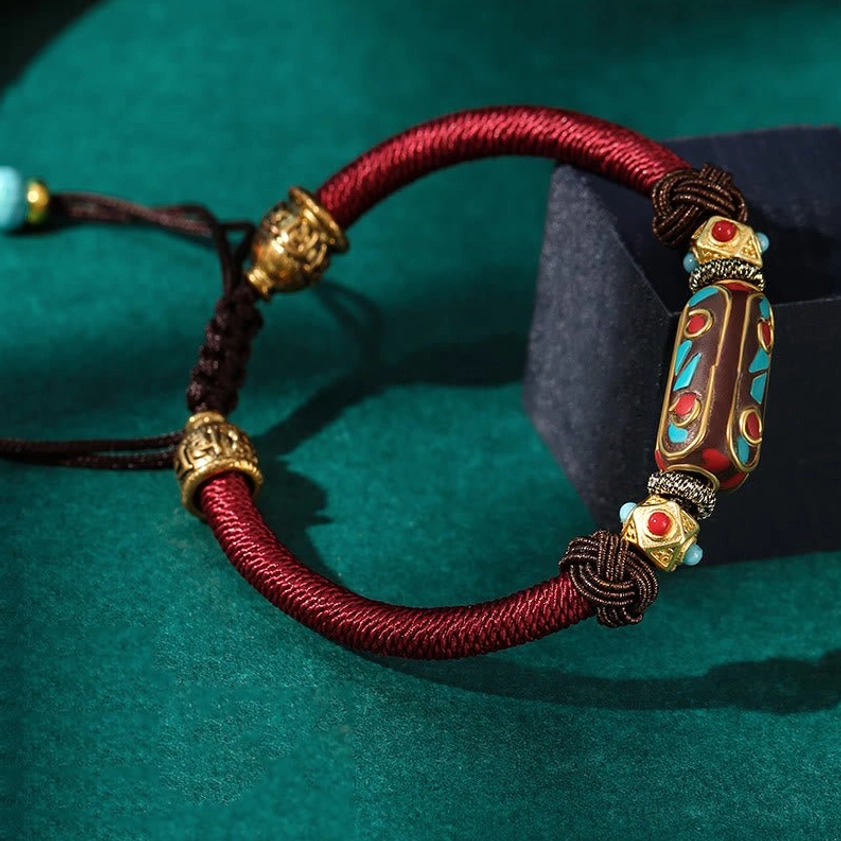 Side profile view of handmade Buddhist bracelet featuring burgundy woven rope with detailed enamel bead, adjustable pull cord and turquoise bead decoration against dark green velvet
