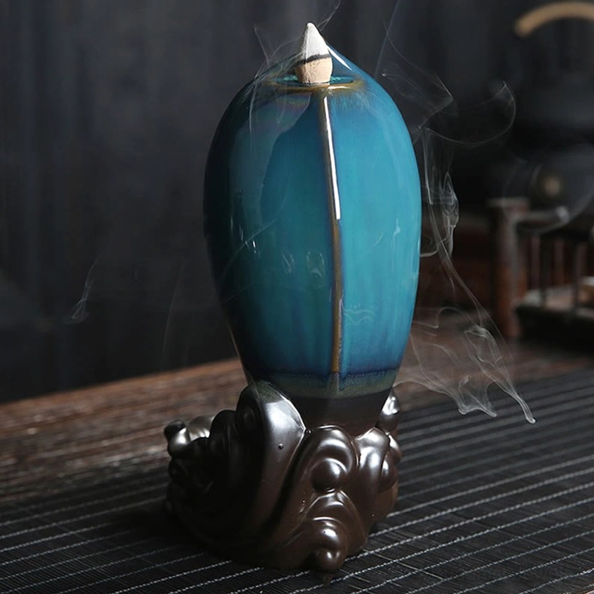 Oval jar shaped backflow incense burner with teal glazed ceramic body and dark brown wave base emitting incense smoke