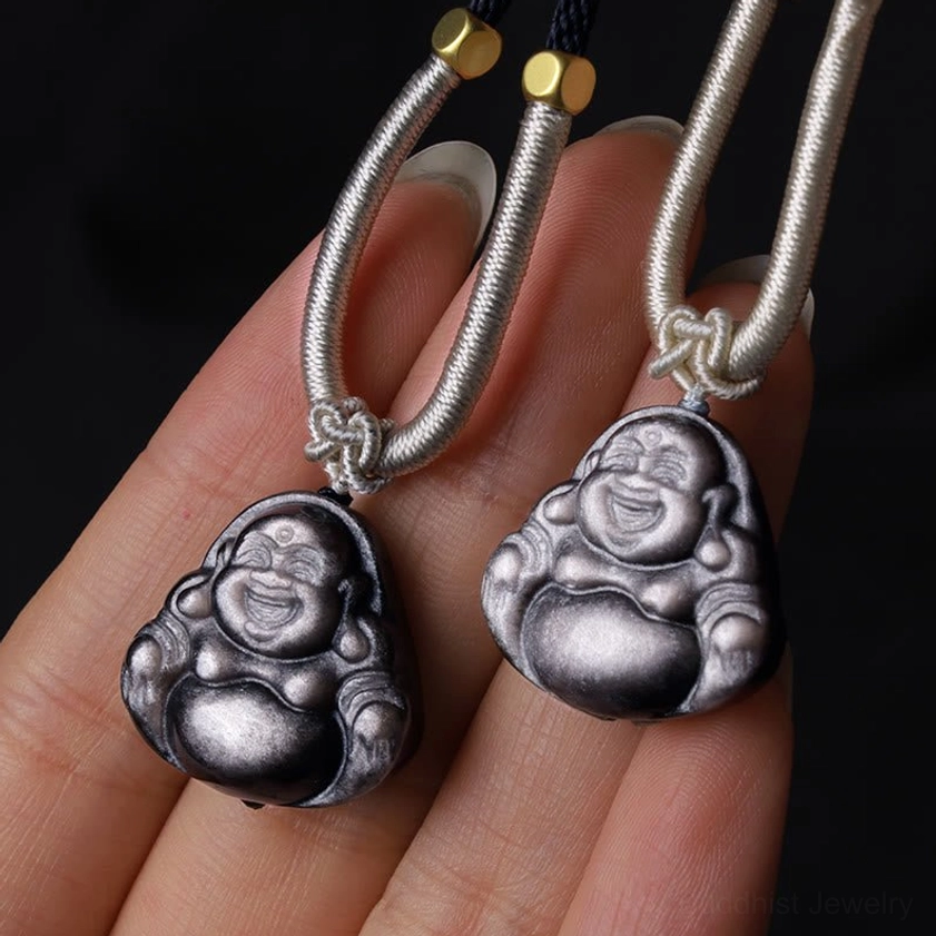 Matching pair of silver sheen obsidian Laughing Buddha pendants on fingers, identical Maitreya Buddha carvings with light grey braided ropes and gold beads
