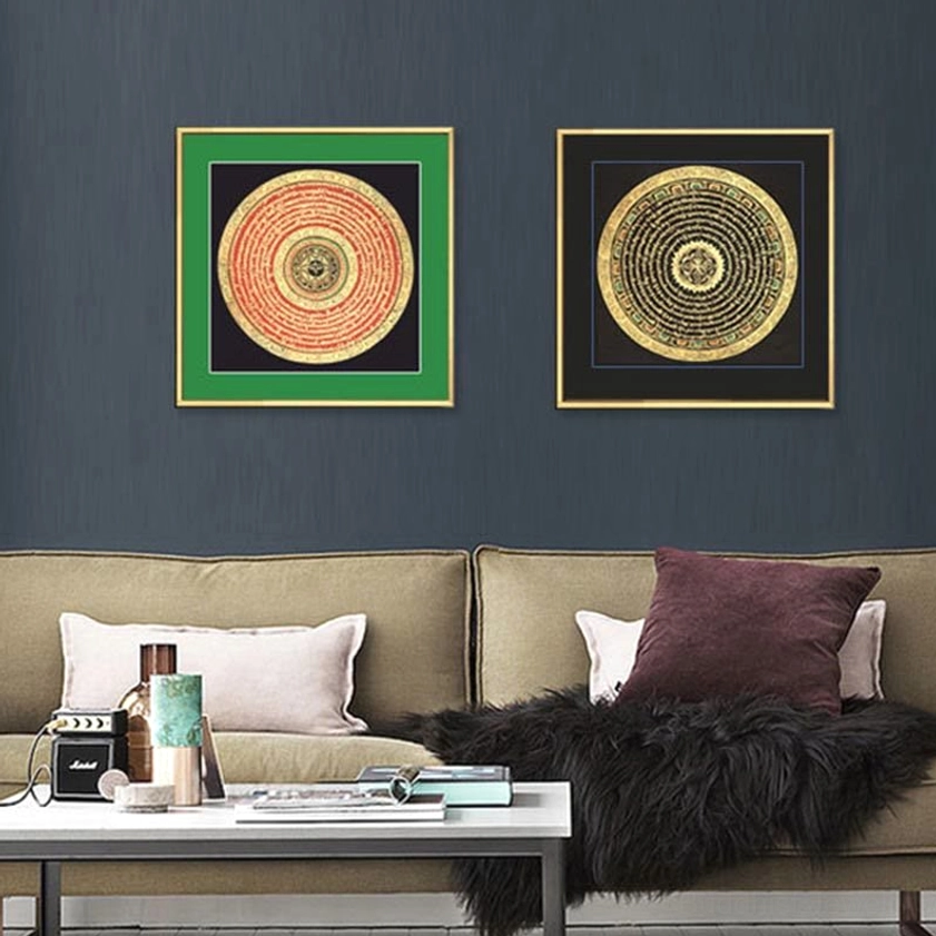 Two mixed Buddhist thangka scrolls displayed above modern sofa in eye-level view, featuring green and dark frames with gilded circular scripture patterns