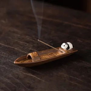 Ceramic panda boat incense burner on dark wood desk with lit incense and rising smoke, side view