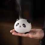 Handcrafted panda ceramic incense burner held in palm, showing matte white finish with black panda design and emitting fragrant incense smoke