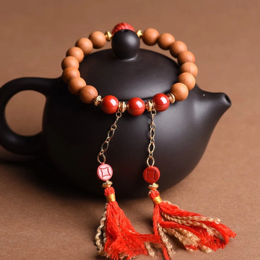 Peach wood prosperity bracelet with dangling red cinnabar tassel charms positioned near black teapot and wooden tray