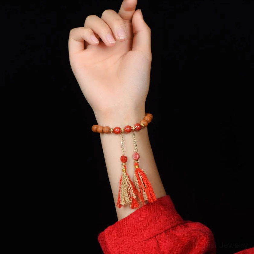 Hand wearing stacked natural peach wood and red cinnabar bracelet with tassel charms against dark backdrop and red clothing