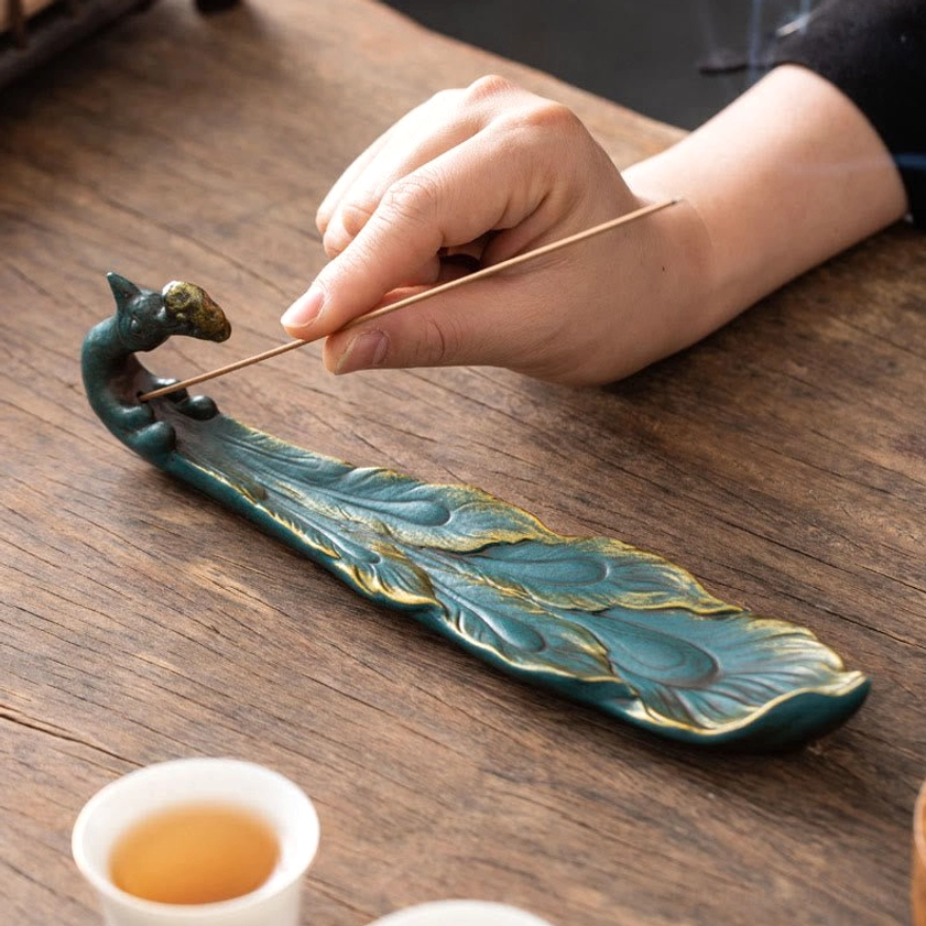 Close-up hand placing incense stick in blue peacock ceramic burner gilded details tea ceremony