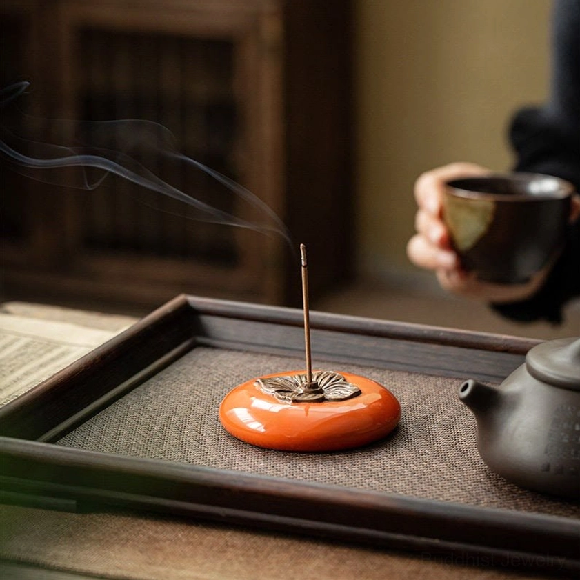 Persimmon incense burner with rising aromatic smoke on tea tray in eye-level close-up, person holding tea cup in background