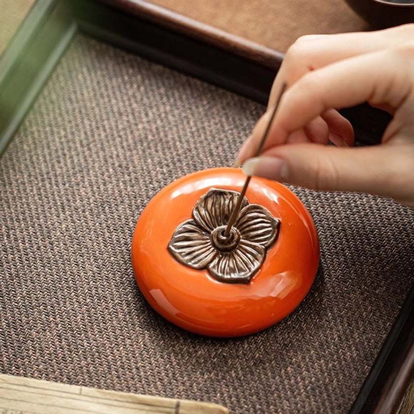 Hand inserting incense stick into ceramic persimmon incense burner on tabletop, showing side angle with bronze decorative details