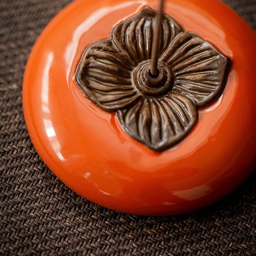 Close-up side view of ceramic persimmon incense holder showing smooth glossy glazed surface, embossed bronze flower pattern, and incense stick