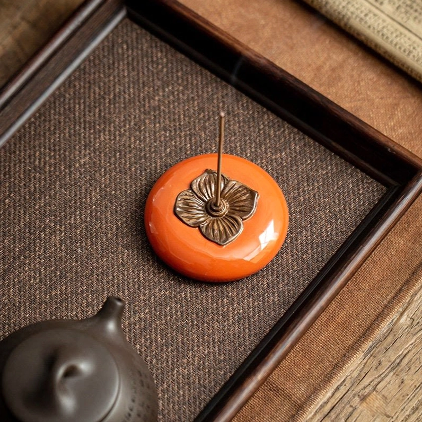 Ceramic persimmon incense holder with inserted incense stick displayed on tea tray next to black teapot in tabletop setting