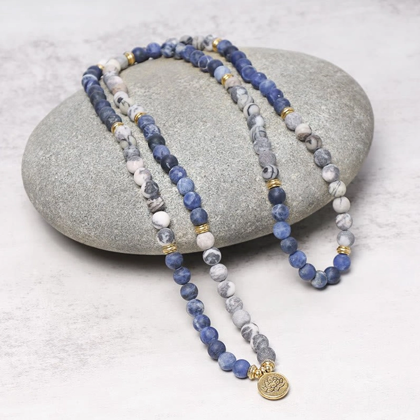 Picasso Jasper 108 bead mala necklace on gray stone surface, showing mixed blue-gray natural stone beads with gold spacers and lotus pendant