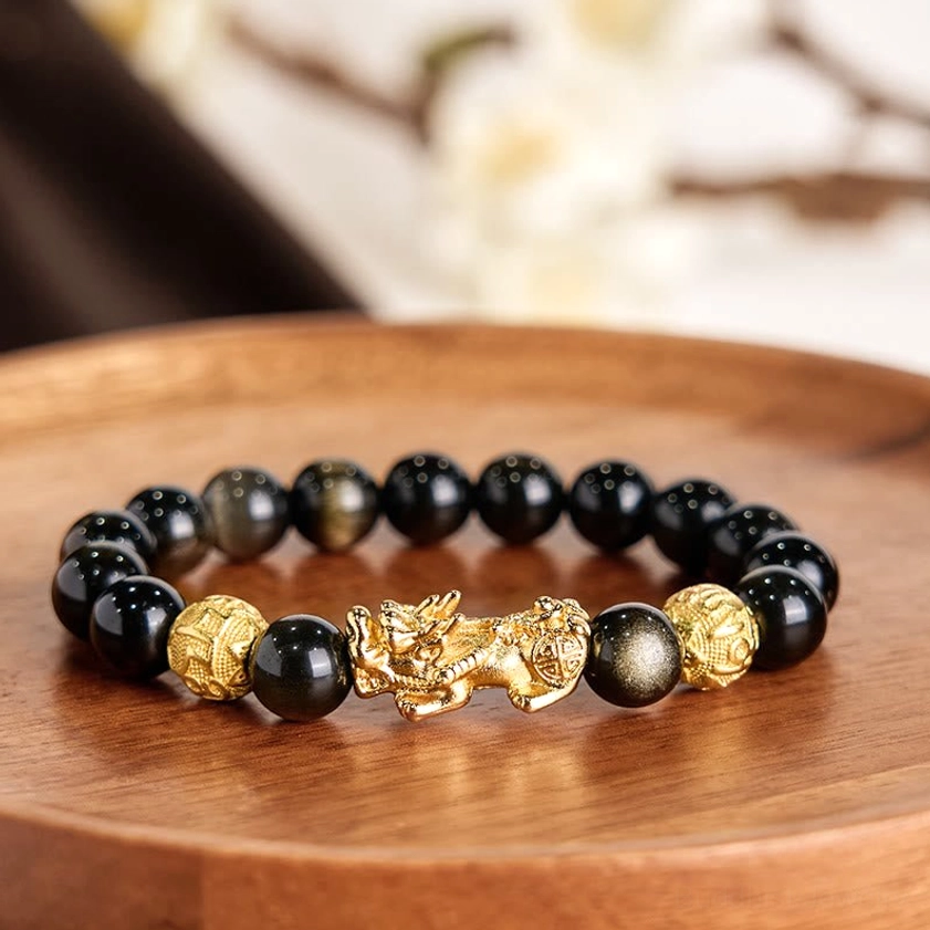 Close-up top-down view of gold sheen obsidian Pixiu bracelet on polished wooden tray, highlighting the central gilded Pixiu figurine and beaded design