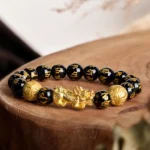 Black obsidian Pixiu wealth bracelet with golden charm on wooden display stand, overhead view showing engraved beads and spacers