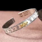 Pixiu silver bangle bracelet with engraved Buddhist Heart Sutra, gold charm ornament, three-quarter view on pink velvet