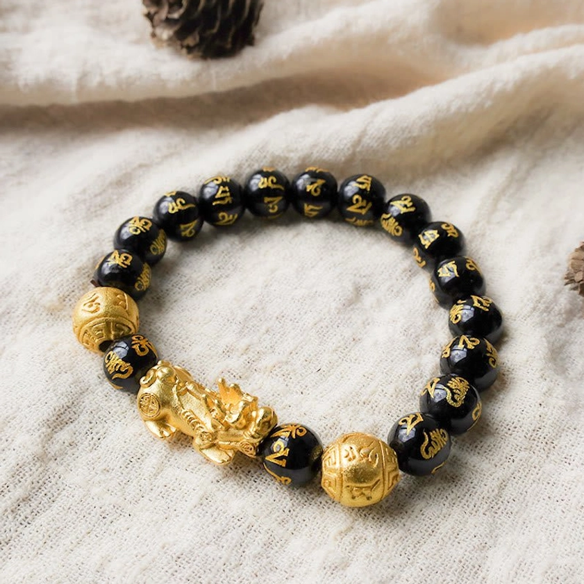 Black obsidian Pixiu wealth bracelet with six true words mantra and golden Pixiu charm, displayed on linen fabric