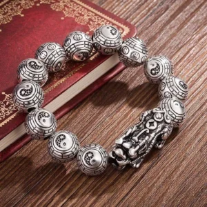 Buddhist Pixiu wealth bracelet with silver carved beads and dragon-lion charm, top-down flat lay on vintage wooden desktop with red book, antique silver finish