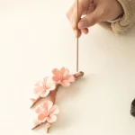 Plum blossom ceramic incense burner with pink flowers on brown branch, incense stick inserted, overhead close-up view