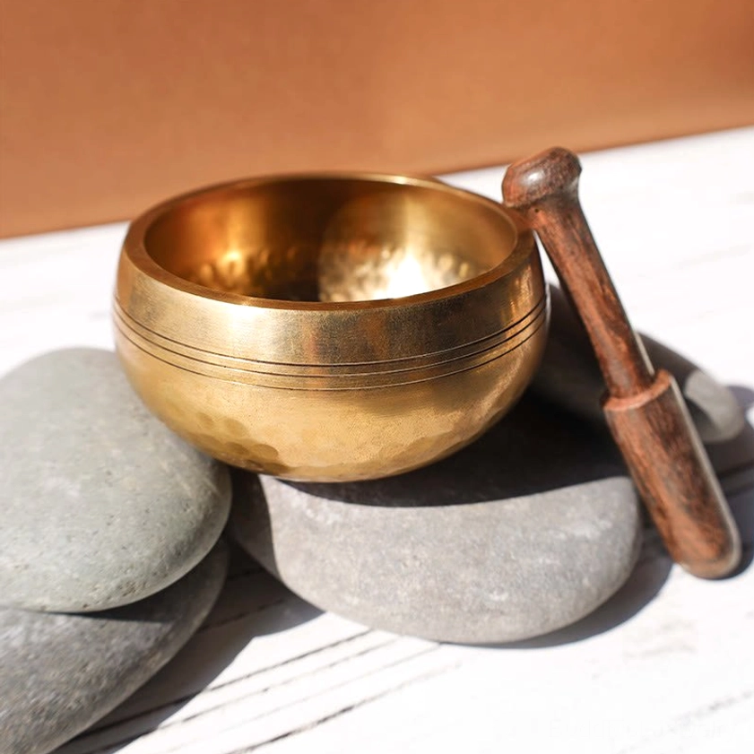 Polished brass Buddhist singing bowl with dark wood striker displayed on river stone surface, demonstrating authentic craftsmanship and natural material finish