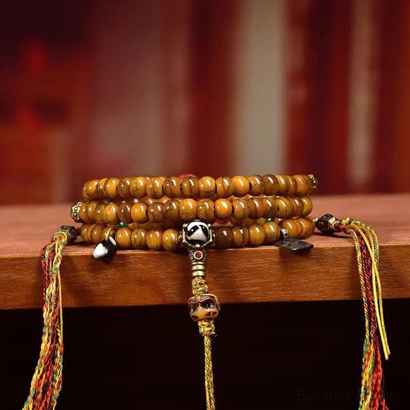 Multiple stacked Buddhist prayer malas with hand-painted Dzi beads displayed on wooden shelf, showing multicolor woven tassels and silver hardware