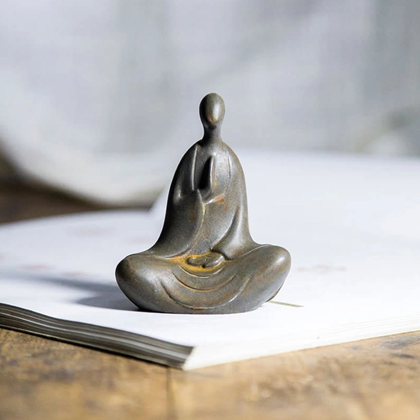 Seated Buddha figurine with aged dark gray finish and prayer pose displayed on wooden desk near closed book