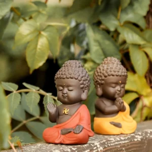Pair of small handcrafted purple clay Buddha statues with red and yellow robes, sitting on wooden surface with green plant background, showing golden swastika marks and serene expressions
